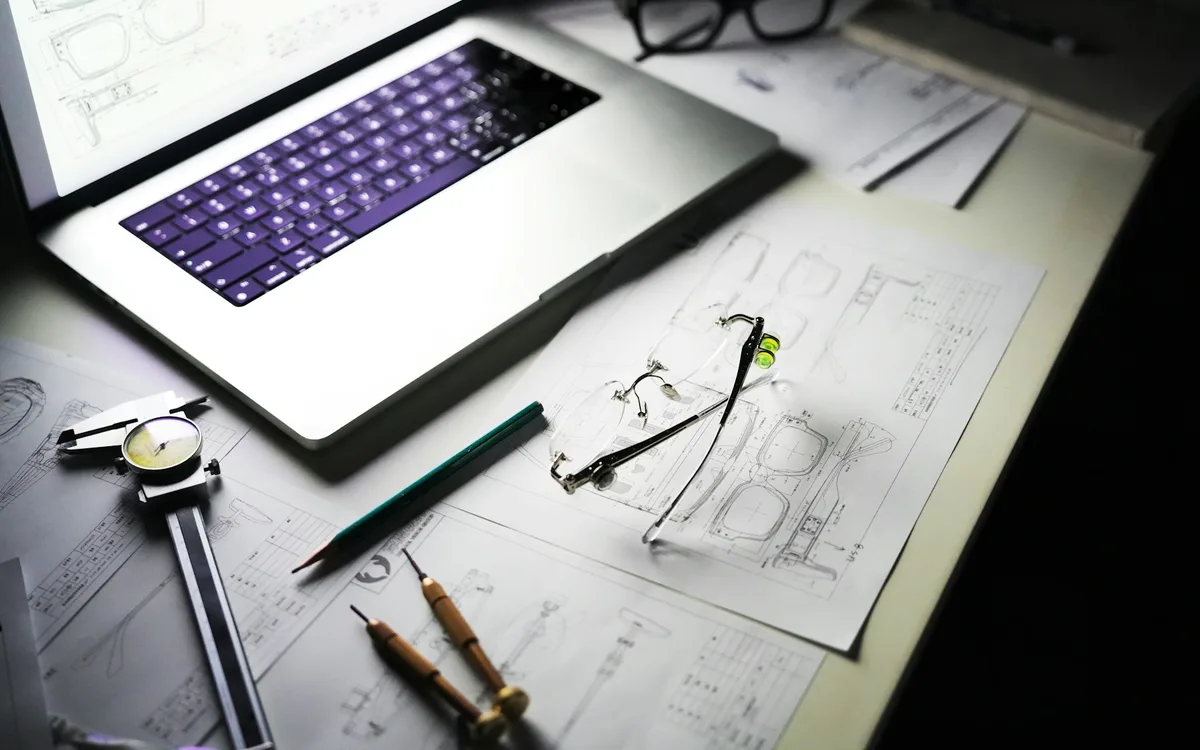 Laptop, rysunki i narzędzia pomiarowe na biurku projektowym.